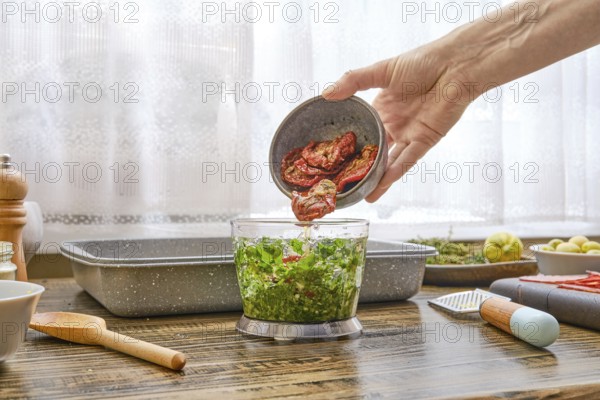 A hand pours dried tomatoes into a bowl of blender filled with vibrant green vegetables. The