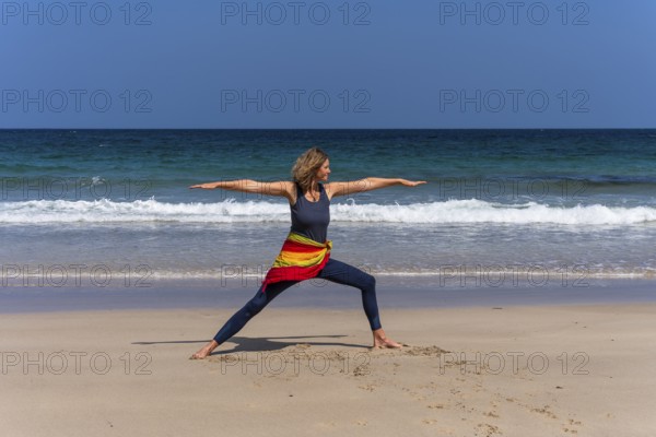 A woman doing yoga on a sandy beach by the sea. Virabhadrasana 2 or Warrior 2. Treen Cove,