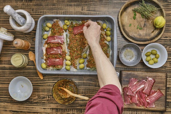 Hand places slices of bacon atop a seasoned fish fillets on a baking tray. Surrounding ingredients