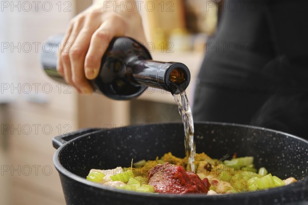 A person pours white wine from a dark bottle into a pot filled with chopped vegetables and