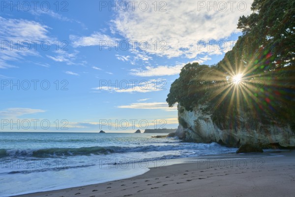 Sandy beach beach with waves and rocks, sun breaking through behind trees in the coastline,