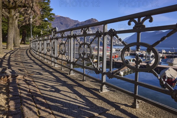 Forged railing with shadow, frog perspective, lakeside promenade, trees, lake, mountains, blue sky,