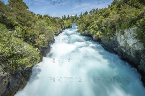 A rushing waterfall at Huka Falls surrounded by lush vegetation and blue sky, Taupo, New Zealand,