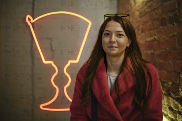 A young woman with long brown hair smiles while wearing a pink coat, seated in a trendy location