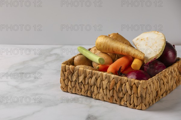 A woven basket full of vegetables such as soup vegetables, potatoes and onions on a marble tabletop