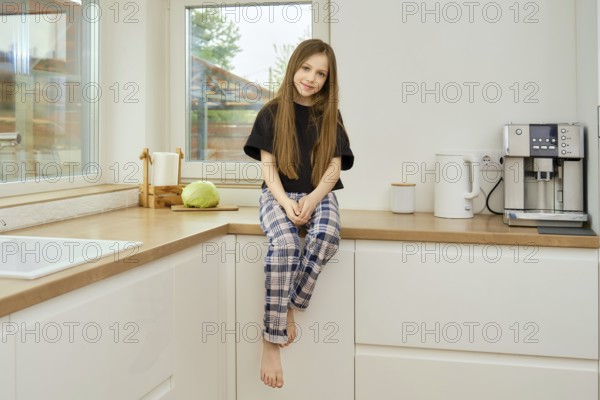 Cheerful young girl in plaid pants and black shirt sits on wooden kitchen tabletop
