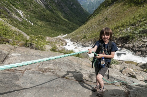 Boy uses rope at steep passage, rock, hiking path to glacier Buarbreen, river in valley stream down