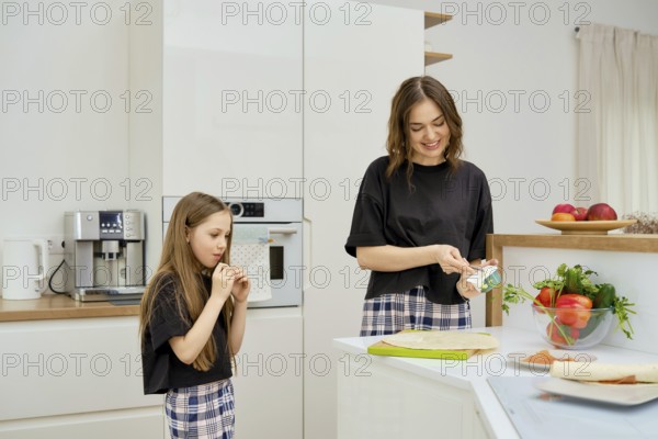 A girl bites a tortilla while her mother spreads soft cheese on the flatbread while preparing a