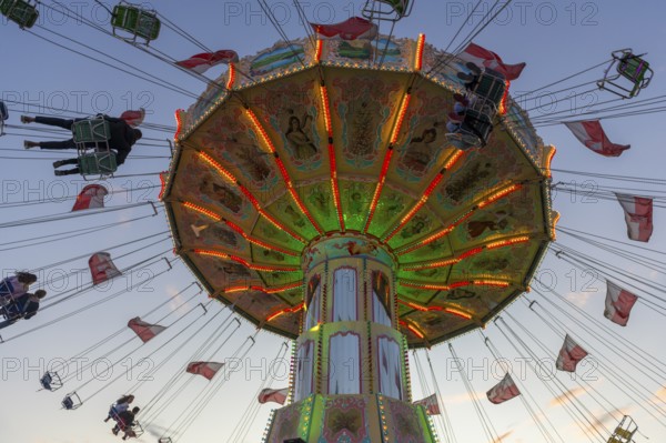 A colourfully illuminated carousel with swinging seats against an evening sky, wave flight, chain