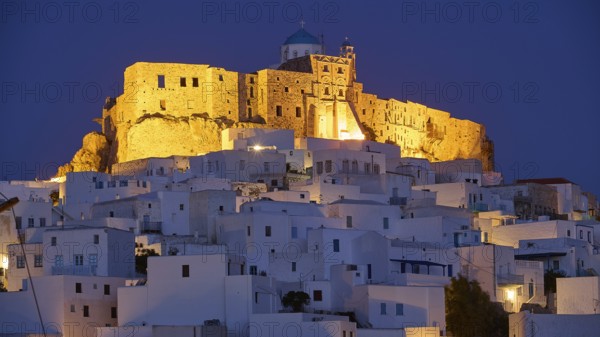 An illuminated historic building towers over plain white structures in the darkness, Querini