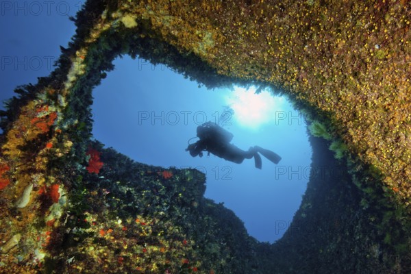 Underwater photo with view through small tunnel breakthrough in rock reef on diver scuba diver in