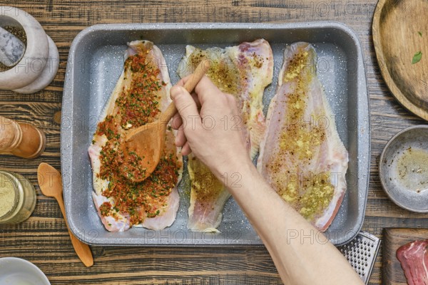 Fish fillets are arranged in a baking tray while a hand spreads a flavorful spice mixture over them