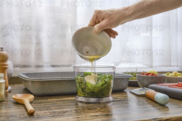Hand pours lemon juice into a food processor filled with fresh green herbs and garlic. The kitchen