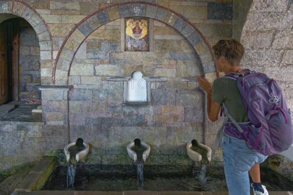 A woman stands in front of the old stone fountain with three water sources and marble basin below