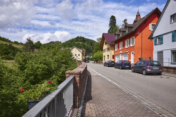 Apartment blocks, hard shoulder with parked cars, hilly landscape with forest, sandstone wall,