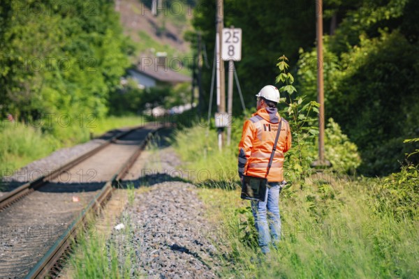 A worker in protective clothing and helmet stands next to railway tracks, surrounded by nature and