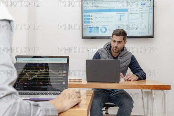 A middle-aged man is deeply focused on his laptop in a modern coworking space, with a screen