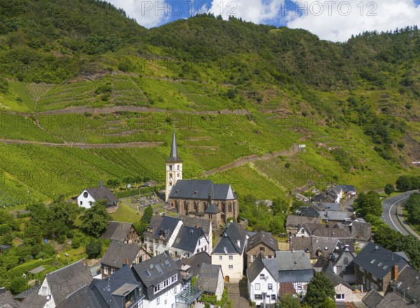 A small village with a church surrounded by vineyards and green hills under a blue sky with clouds,