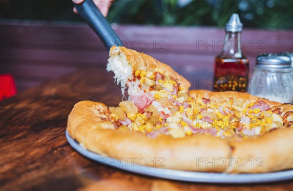 Person hand taking a slice of sweetcorn and ham pizza with spatula. Delicious ham pizza with cheese