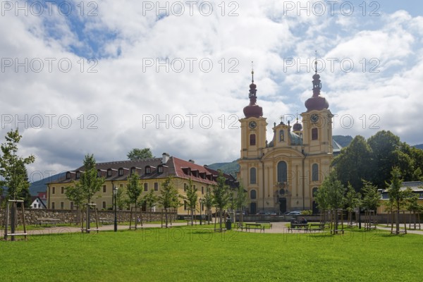 Baroque church with red roofs, surrounded by trees and green meadow with cloudy sky, monastery and