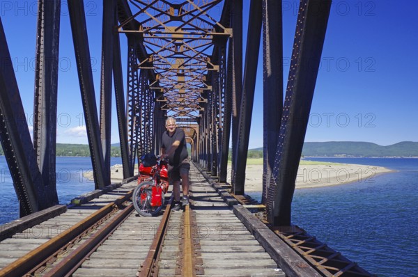 A railway bridge crossed by a person on a bicycle over the sea, surrounded by nature and blue