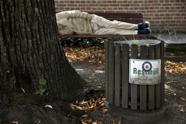 Homeless man lying on a bench in front of a rubbish bin labelled Restmüll, Alter cemetery, Bonn,