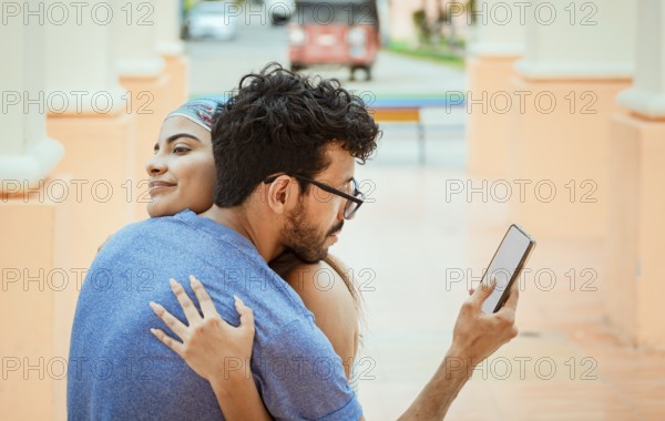 Unfaithful man looking at the cell phone while hugging his girlfriend outdoors. Unfaithful