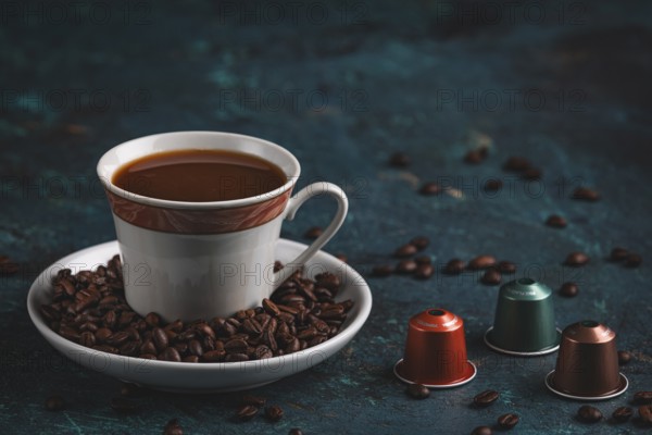 A cup of coffee next to three coffee capsules, surrounded by coffee beans on a textured surface