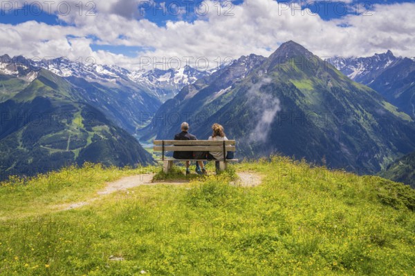 Two people sit on a bench and look out over a valley with majestic mountains in the background,