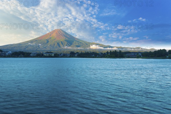 Mount Fuji bathes in morning side light while Lake Kawaguchiko sits in shadow below cloudy blue sky