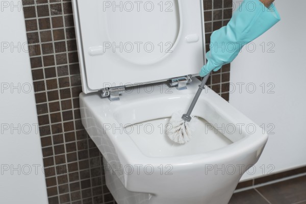Person wearing a turquoise rubber glove cleaning under the rim of a toilet bowl with a toilet brush