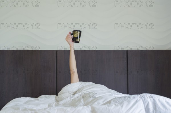 Drinking coffee early tomorrow. Woman in bed, under duvet, with arm raised up holding coffee cup