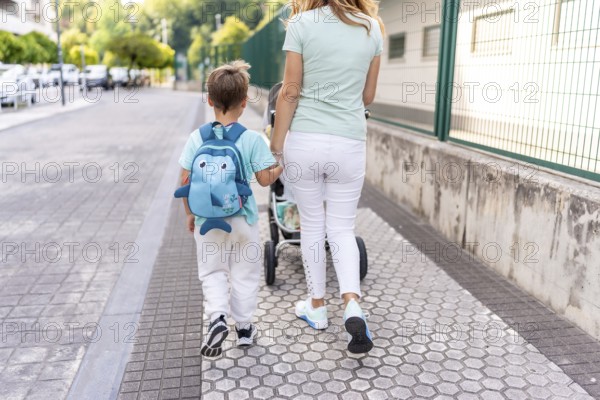 Rear view of a mother and son carrying baby carriage walking to the school together