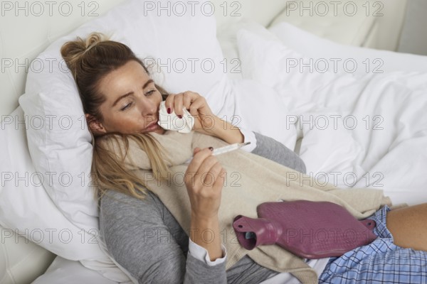Sick woman with a miserable expression taking her temperature as she lies in bed suffering from a