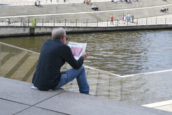 A man sits on a step in the government district of Berlin and reads the financial section of a