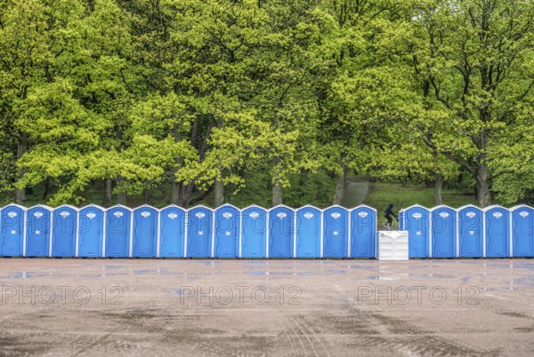 Long row of portable toilets in front of a forest where one has tipped over