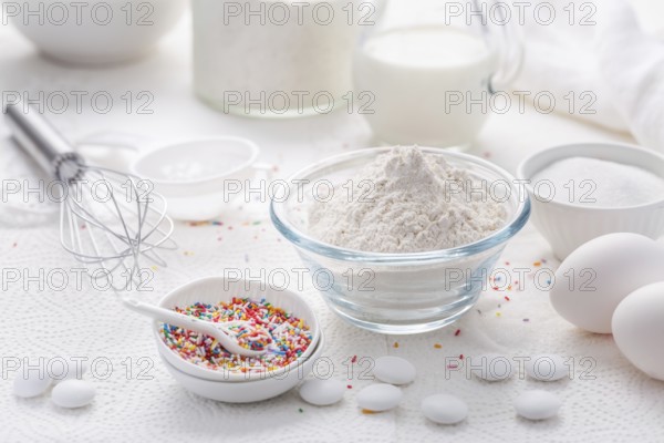 Baking background with flour, eggs, sugar sprinkles, kitchen tools, utensils in white