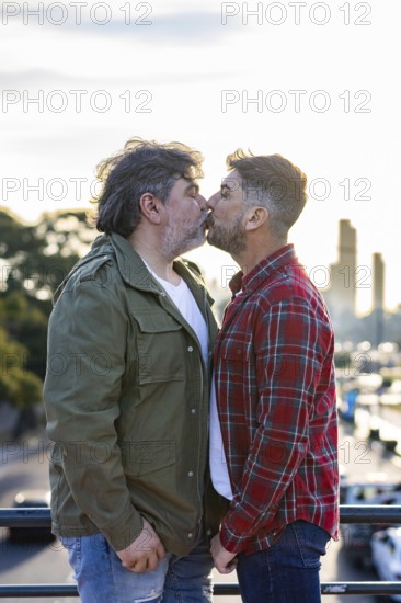 A gay couple, is standing on a city bridge while kissing each other