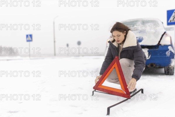 Woman puts up a warning triangle on a snowy road in winter, car breakdown in winter