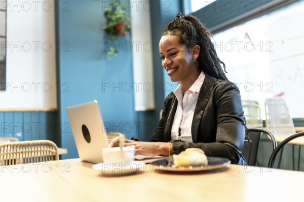 Confident latin businesswoman working with laptop in a cafeteria while eating sandwich and drinking