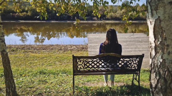 Single woman sitting on the bench at the lake surrounded by autumn foliage, beneath birch trees in