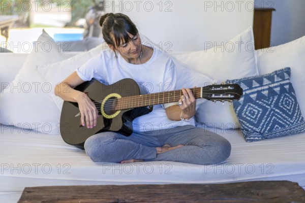 Adult woman playing the guitar while seated on her sofa at home