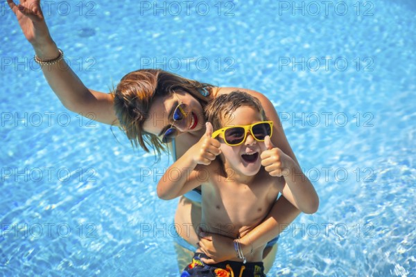 Caucasian boy expressing that he is having fun with his mother in the swimming pool raising thumbs