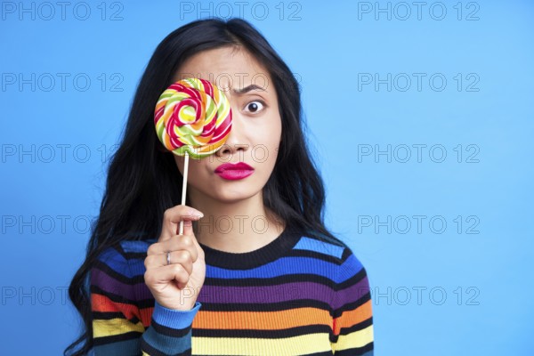 Confused thoughtful woman covers her eye with colorful lollipop looking to camera isolated on blue