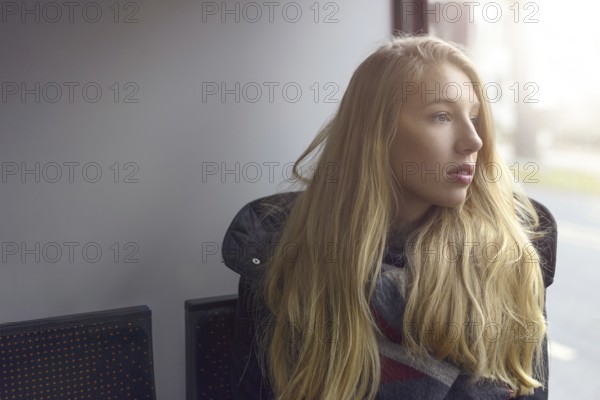Young blond woman sitting in shadowed natural light looking out of a windows at the street outside