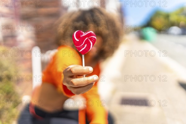 Focus on the hand of a young latin woman holding a heart shape lollipop in the street