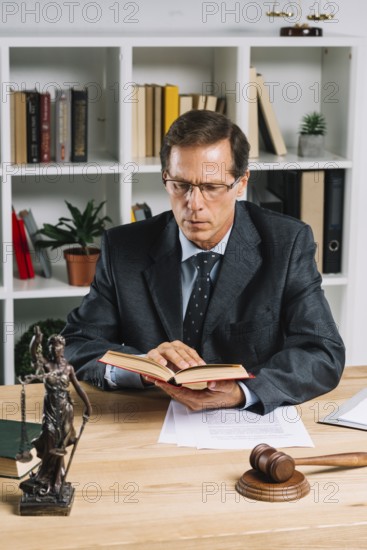 Mature male lawyer reading book with gavel justice statue wooden table