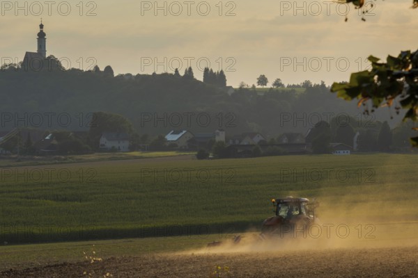 Typical Bavarian rural farm labor scene with tractor plowing field and pilgrimage church on top of