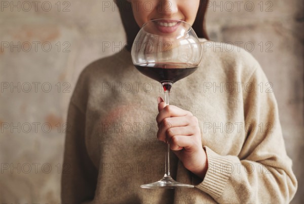 Crop happy female in sweater smelling red wine in large glass against gray background