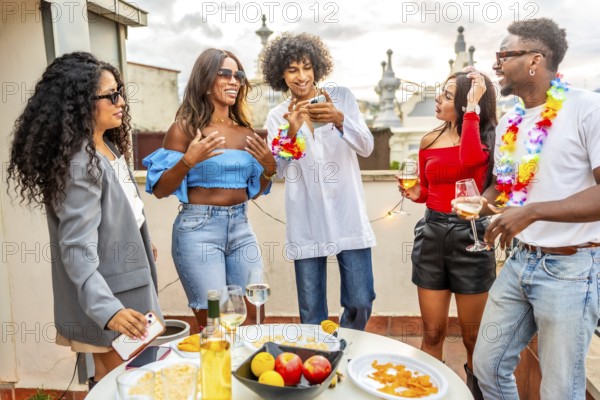 Multi-ethnic friends using mobile phone during evening rooftop party with snacks and drinks